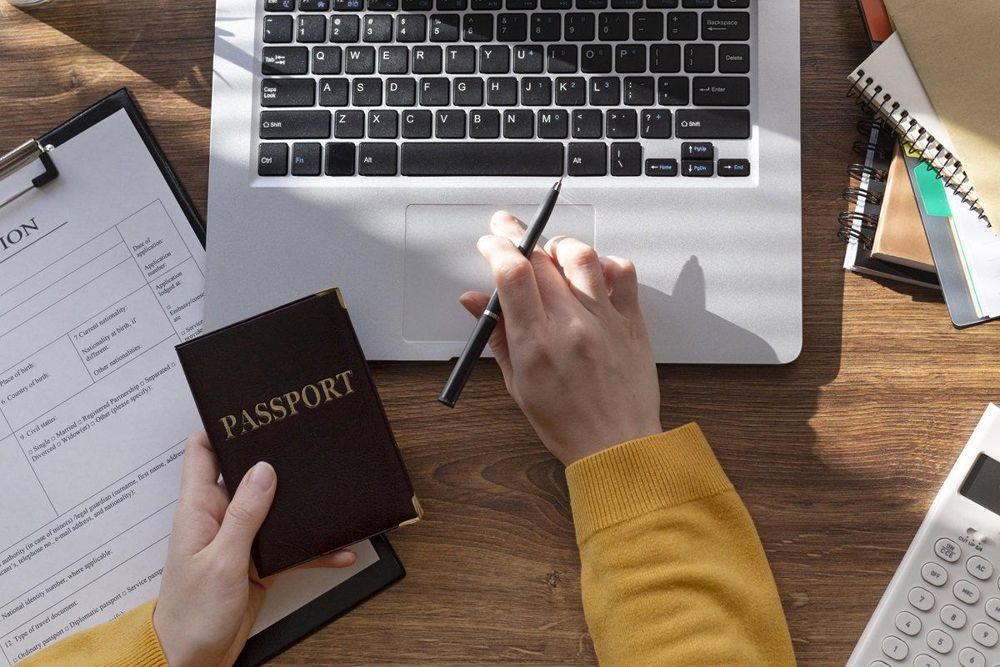A person holding a passport above an application form, with a laptop, notebooks and a calculator on a desk.
