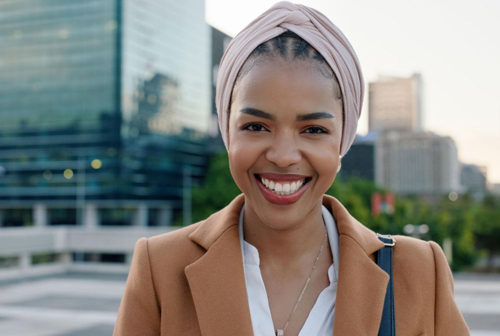 A smiling woman in a camel coat and pink head wrap standing in a city.
